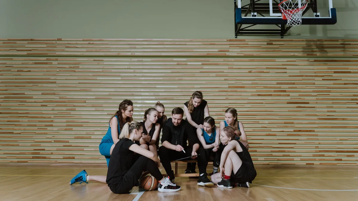 A group of young women in basketball uniforms are gathered around a man in a black shirt, who appears to be their coach or trainer, on a bas