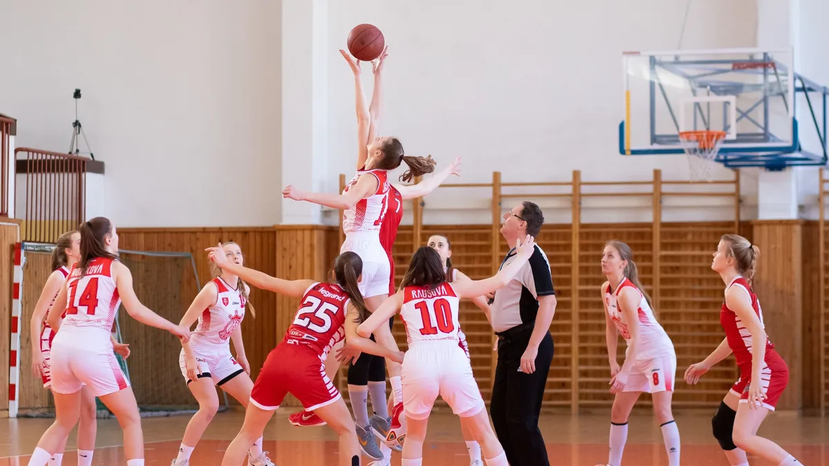 A group of young women in red and white uniforms are engaged in a basketball game, with one player attempting to shoot the ball into the hoop.