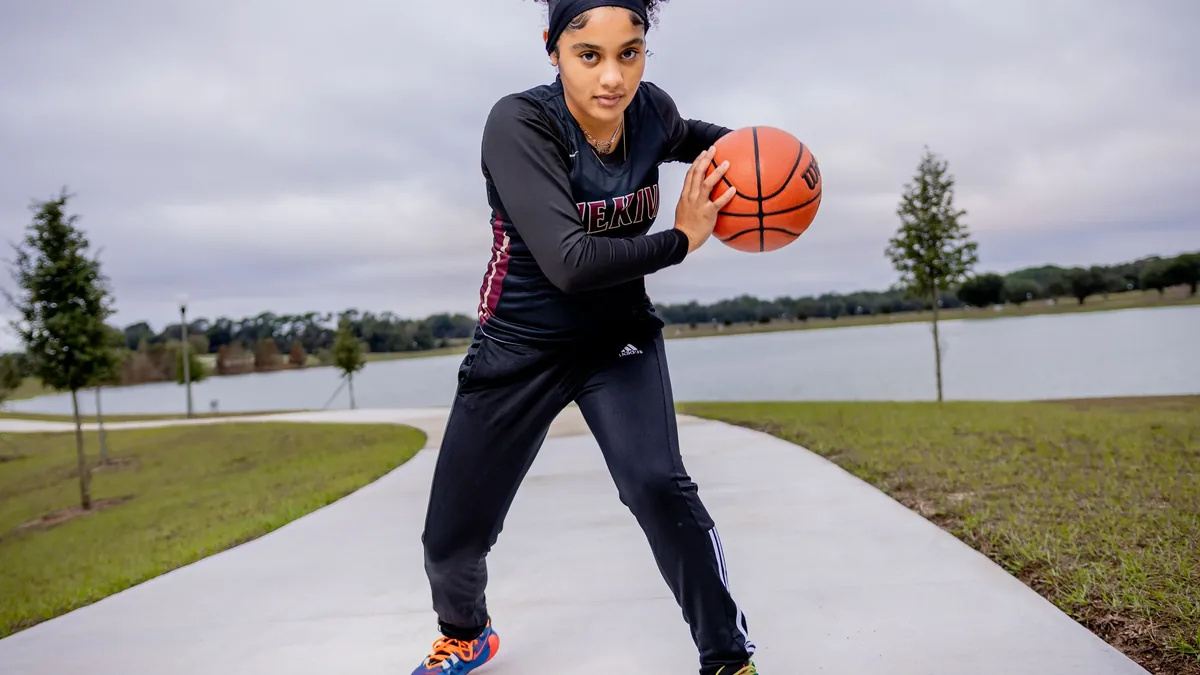 A young woman in athletic attire, holding a basketball and standing on a sidewalk.