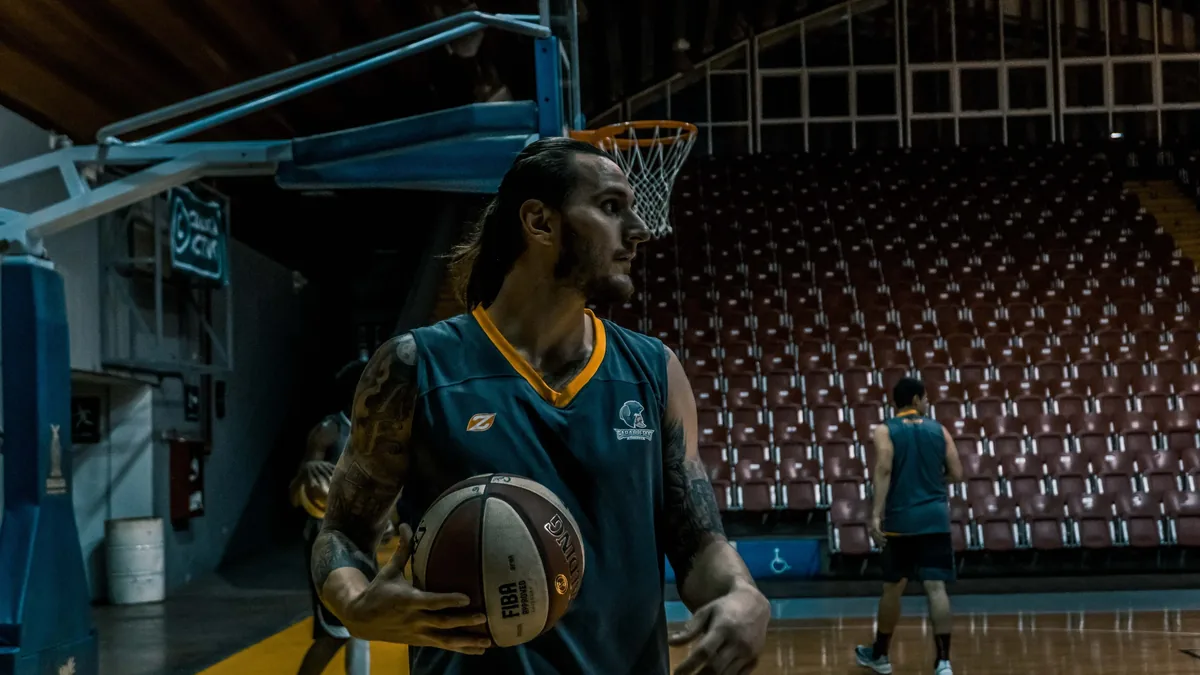 A man with tattoos is standing in a gymnasium, holding a basketball and wearing a blue jersey with yellow trim.
