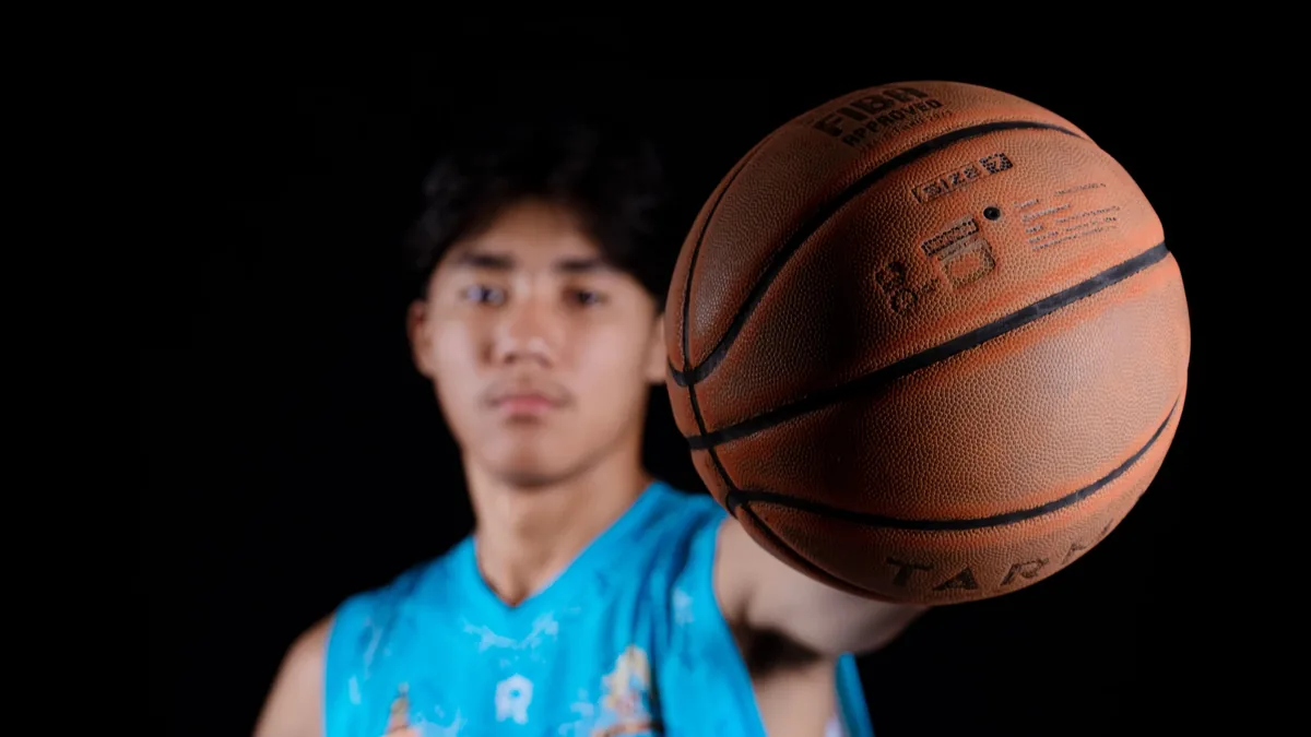 A young man is visible from the chest up, wearing a blue sleeveless jersey with an indistinct design, and is holding a basketball in his rig