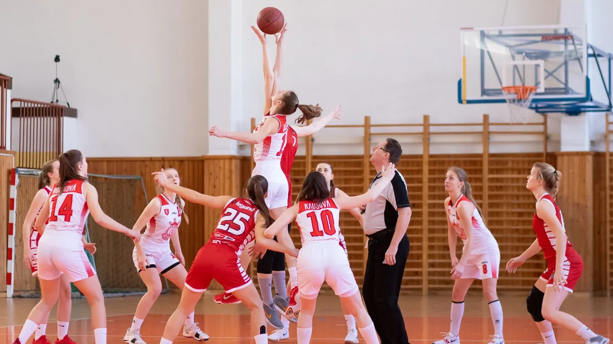 A group of young women in red and white uniforms are engaged in a basketball game, with one player attempting to shoot the ball into the hoo